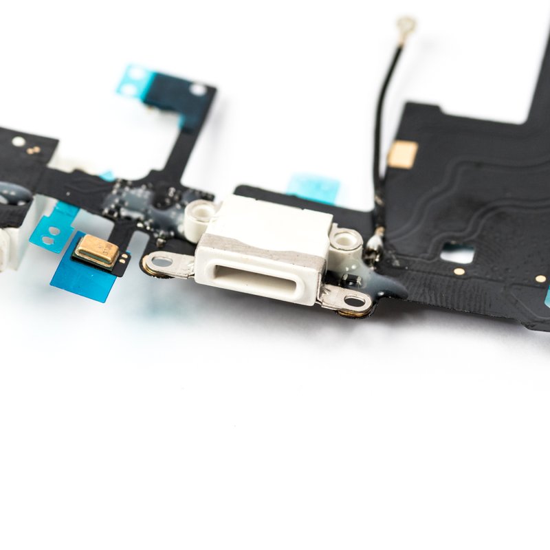 Close macro of a white iPhone 5 charger flex, focused on the white Lightning connector with its metal screw tabs, microphone and teal protective tabs on black ribbon.