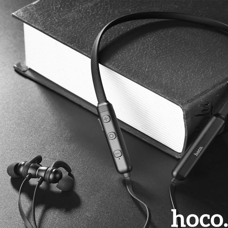 Black and white photo of the HOCO ES11 Maret Bluetooth neckband earphones draped over a book, showing the inline control buttons and ear hook earbuds, hoco logo.